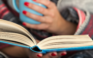 A woman wrapped in a blanket reads a book while holding a mug