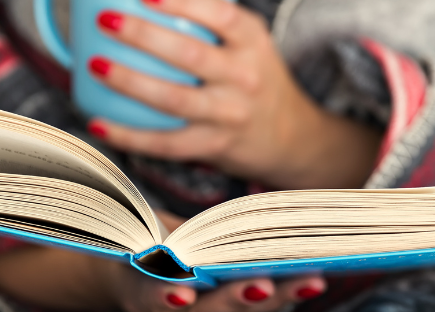 A woman wrapped in a blanket reads a book while holding a mug