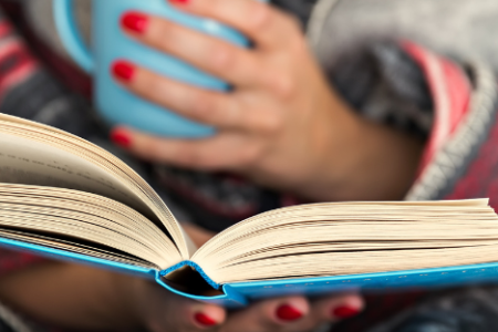 A woman wrapped in a blanket reads a book while holding a mug