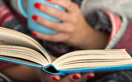A woman wrapped in a blanket reads a book while holding a mug