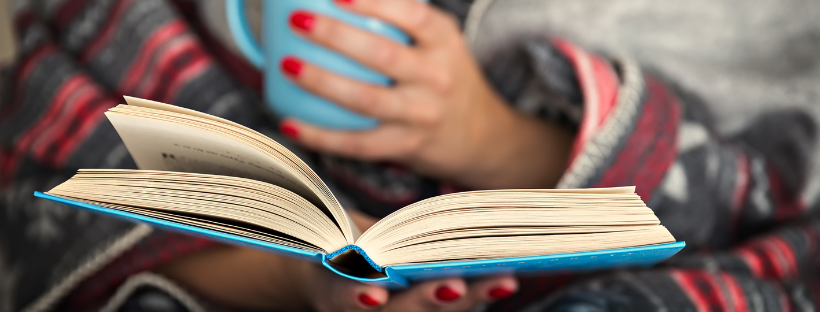 A woman wrapped in a blanket reads a book while holding a mug